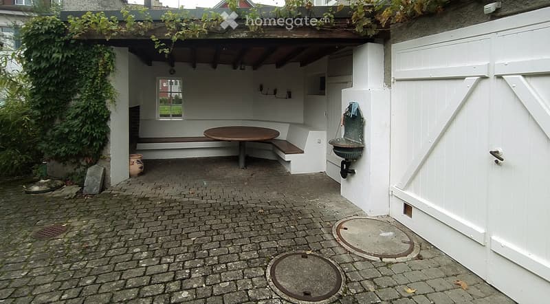 A small outdoor patio with a wooden table, bench seating, a decorative fountain, and a covered roof supported by wooden beams.