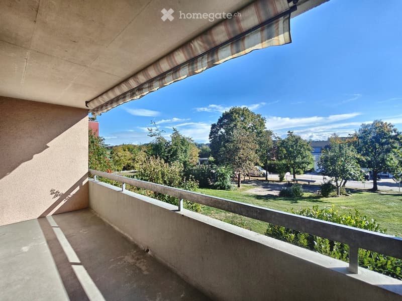 Balcony with a striped awning, overlooking a green park area with trees, grass, and a few benches.
