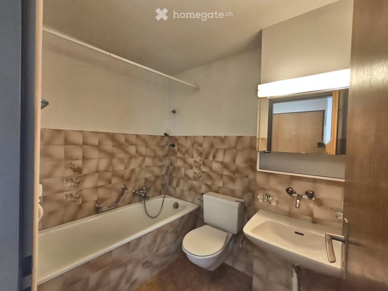 Modern bathroom with a bathtub, toilet, sink, and mirror. Features beige tiled walls and a built-in cabinet with lighting above the mirror.