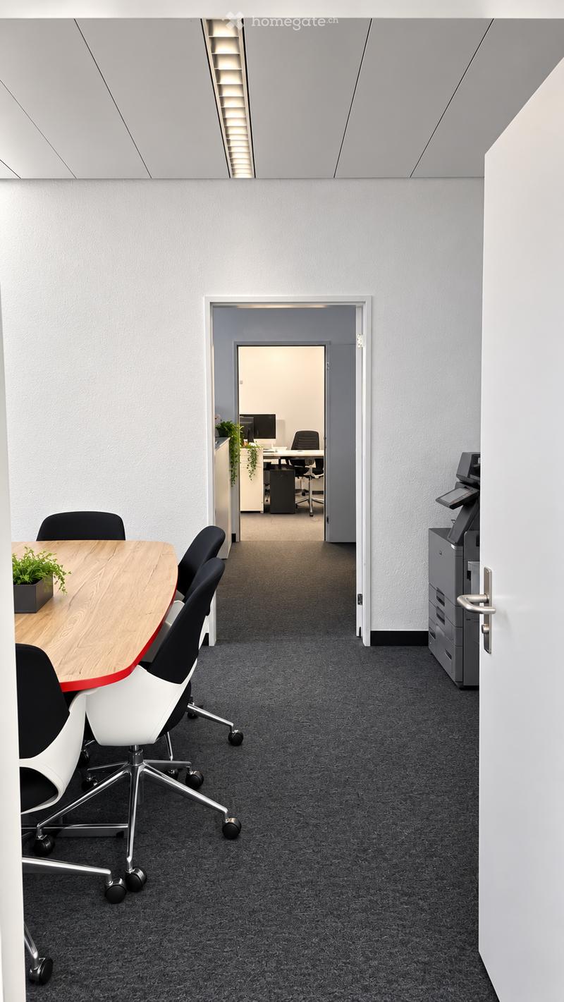 Modern office corridor with grey carpet, white walls, and a wooden table with chairs in the foreground. A printer and filing cabinet are visible to the right, and an open doorway leads to another office space with desks, chairs, and a computer.