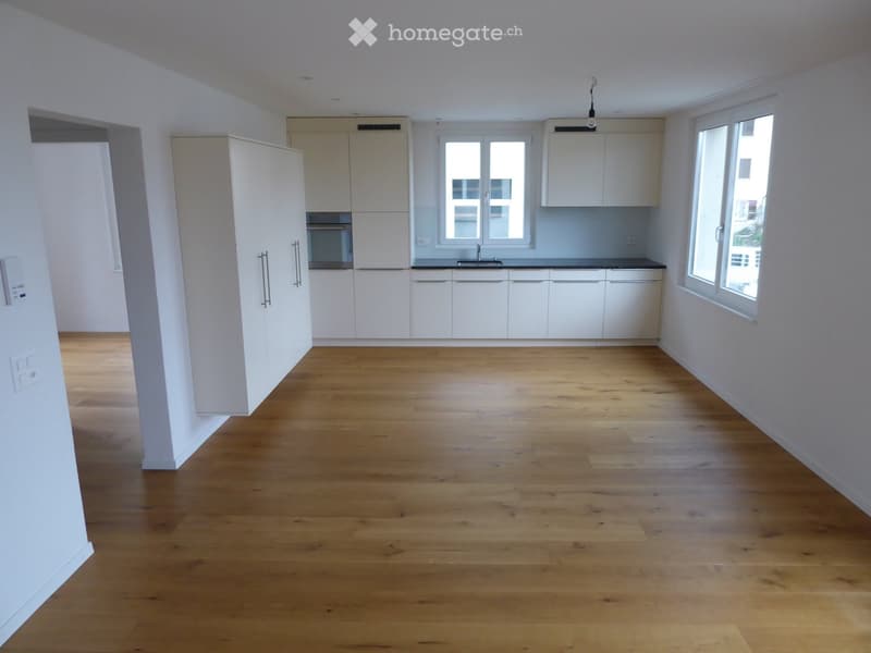 Empty kitchen with white cabinets, wooden flooring, and large windows allowing natural light.