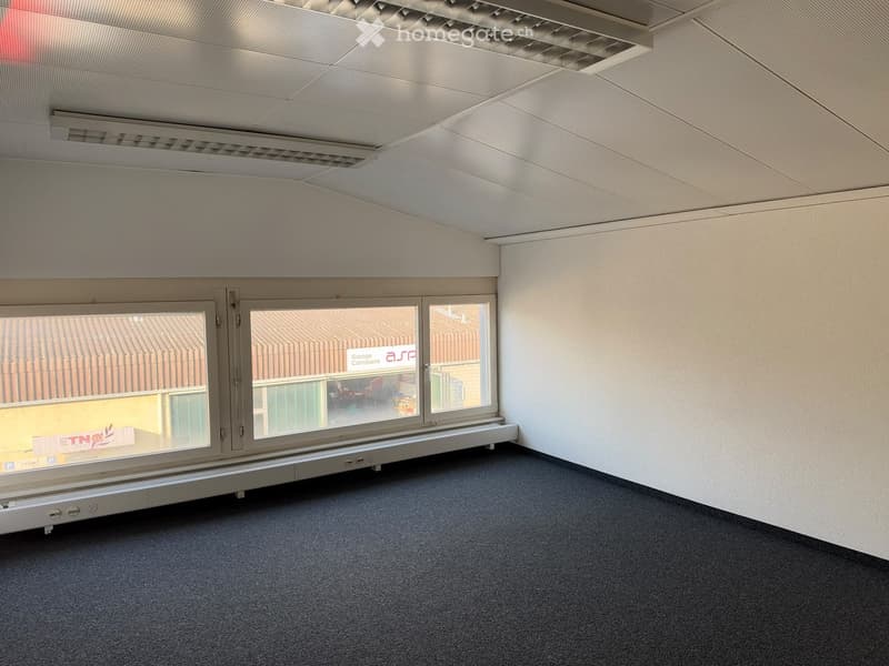 Empty room with grey carpeted floor, white walls, large window with metal frame, and a white ceiling with ventilation grilles.