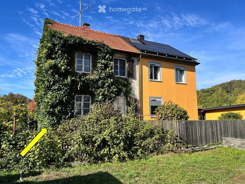 Two-story house with yellow siding, red-tiled roof, and solar panels on the roof. The left side is covered in ivy, and the right side has multiple windows and a small balcony. A wooden fence and greenery surround the house.