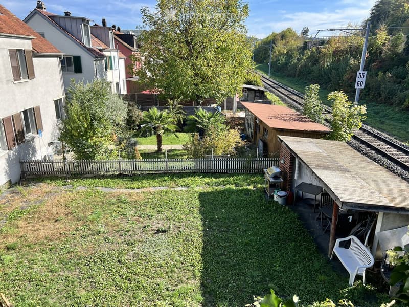 A small, grassy garden area with a wooden fence, a few trees, a small shed, and outdoor furniture including a white plastic chair and a table.