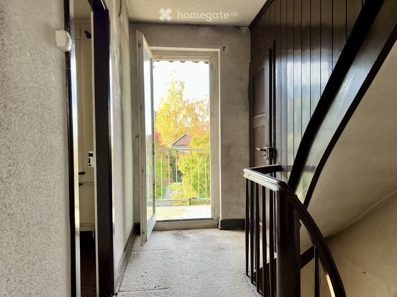 Hallway with carpeted floor, open door leading to a balcony with outdoor view, staircase with wooden railing on the right side.