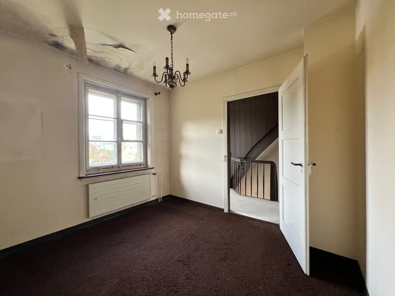 Empty room with beige walls, brown carpet, a window with white frame, radiator, and an open door leading to a staircase.