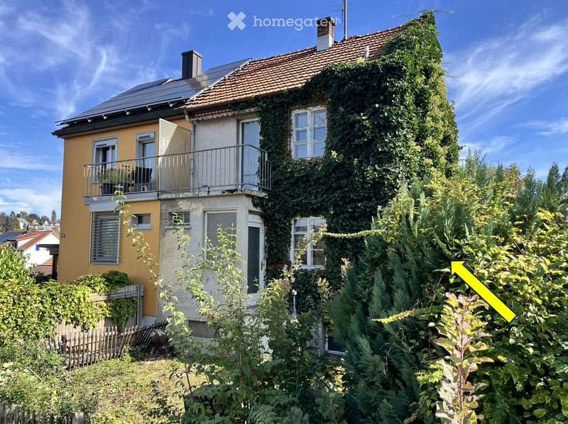 Two-story house with yellow and beige exterior, covered in ivy, featuring a balcony, multiple windows, and a small garden area.