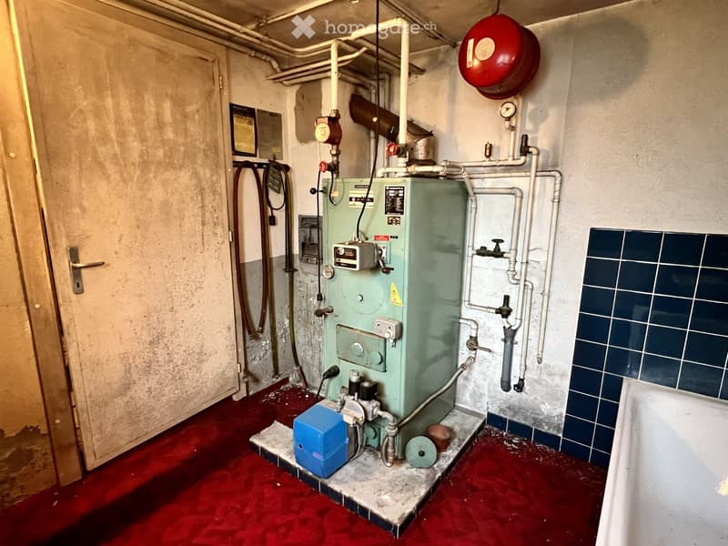 Utility room with heating system, pipes, and a red fire alarm on the wall. The floor is covered in red material, and there is a door on the left side.