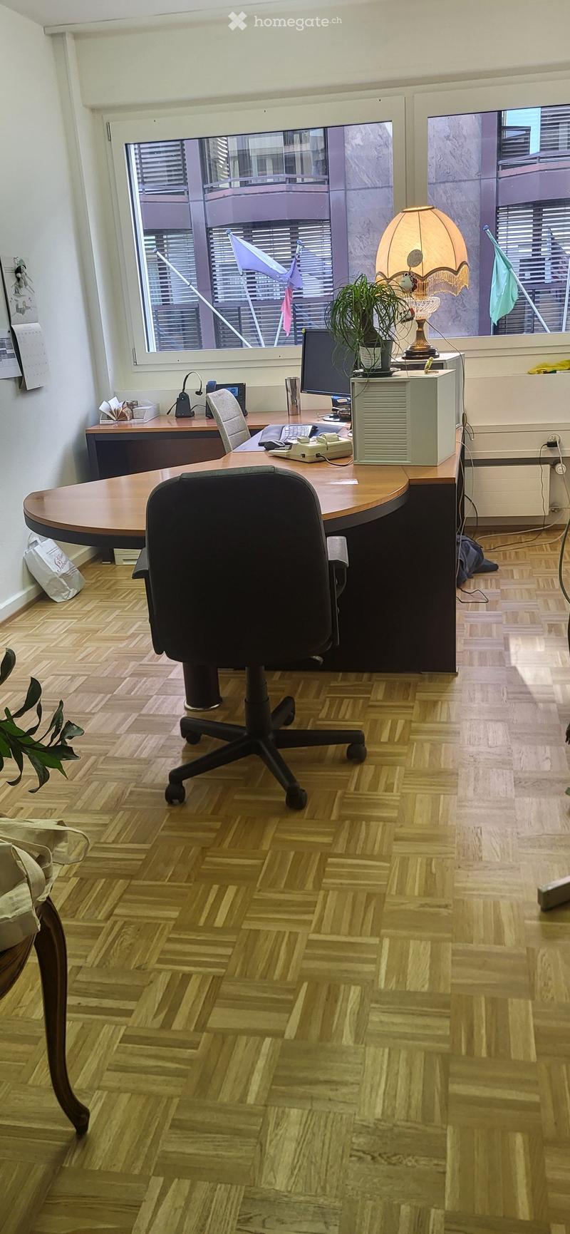 Office space with a wooden herringbone floor, large window with blinds, and a modern office setup including a desk, office chair, computer monitor, lamp, and telephone.