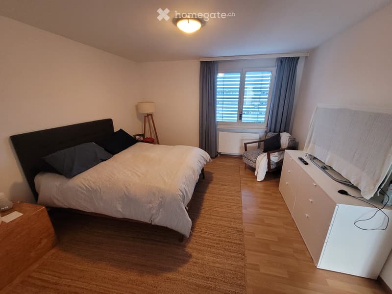 Cozy bedroom with a large bed, nightstand, armchair, dresser, and a lamp. The room features wooden flooring and large windows with blinds.