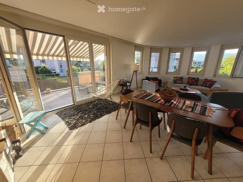 Bright living room with large windows and a balcony, featuring a sofa, dining table, chairs, and a rug.