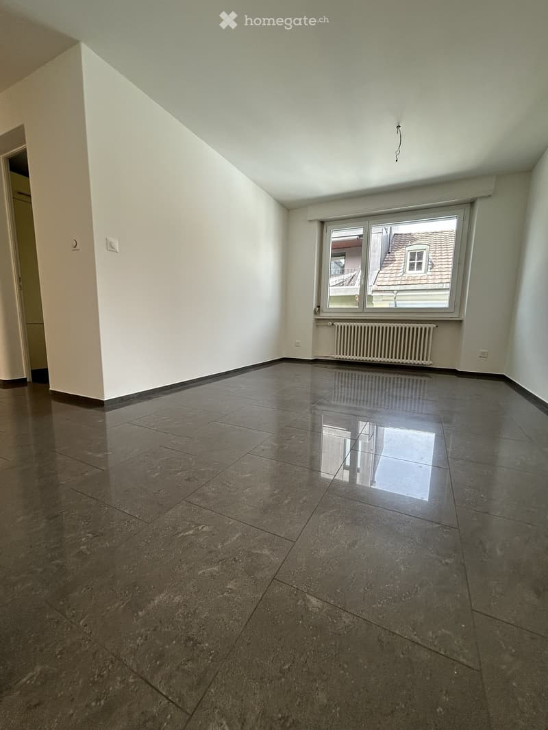 Empty room with polished tile flooring, white walls, and a large window with a view of a roof and neighboring building. A radiator is visible beneath the window.