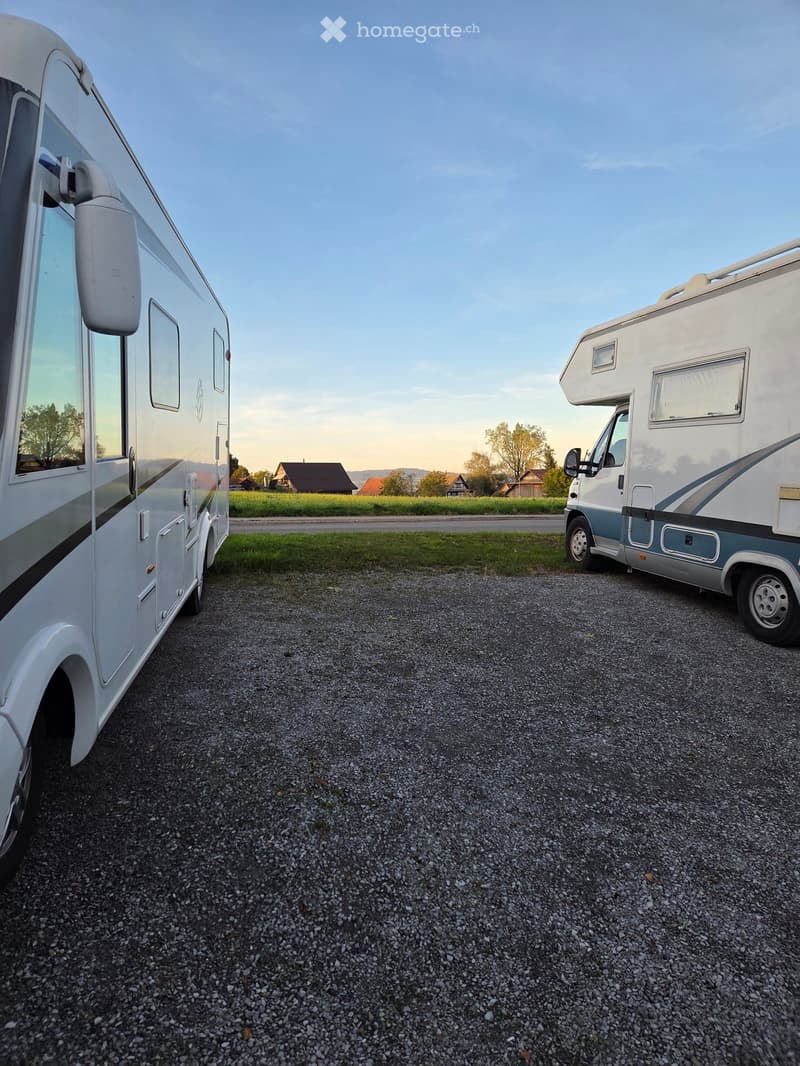 Two RVs parked on a gravel surface with a rural landscape in the background, featuring houses and trees.