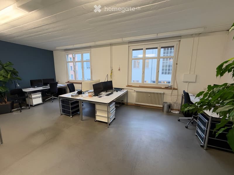 Office space with desks, chairs, computer monitors, and plants, featuring large windows and exposed pipes on the ceiling.