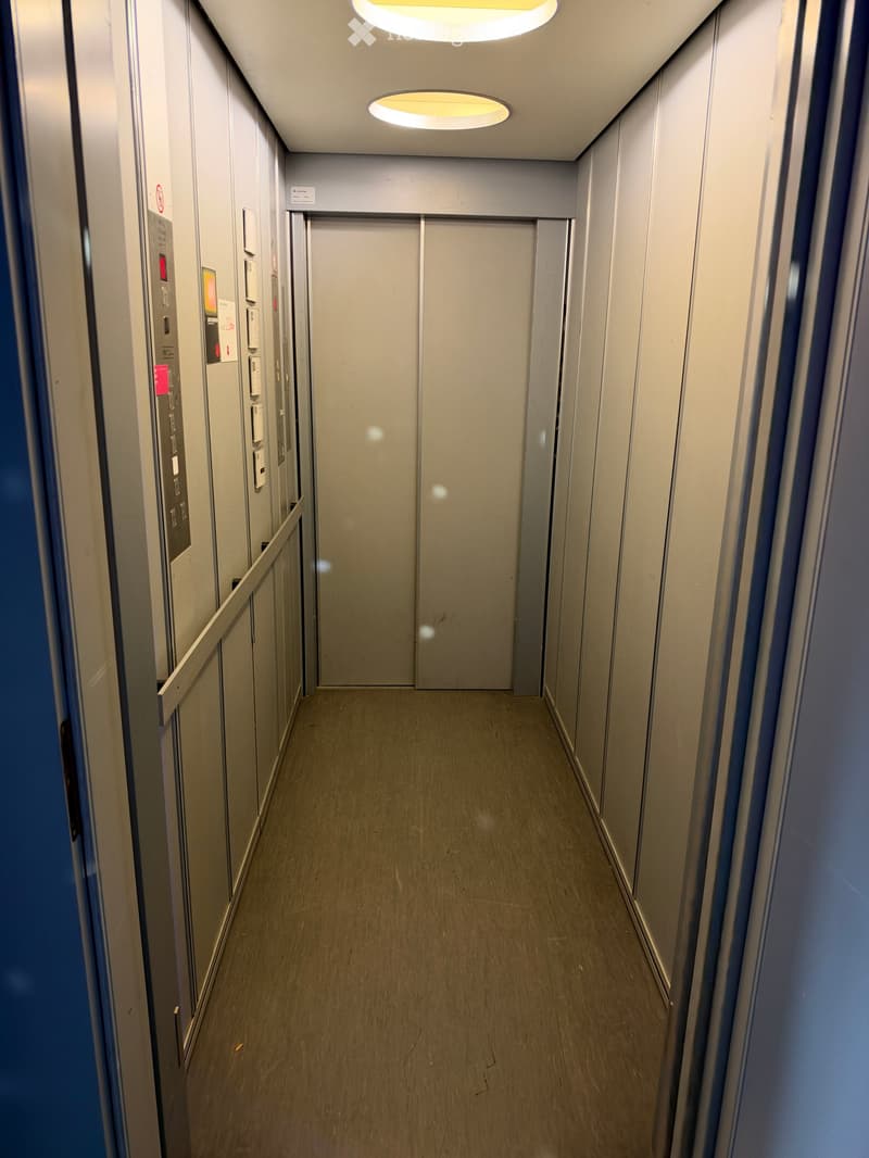 Modern hallway with wooden flooring, grey walls, and recessed lighting. Features elevator doors and control panel on the left side.