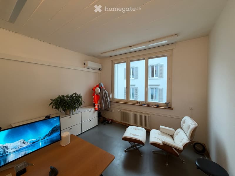 A modern, well-lit room with a large window, a wooden table with a screen, two white chairs, a small plant, and a cabinet with drawers.
