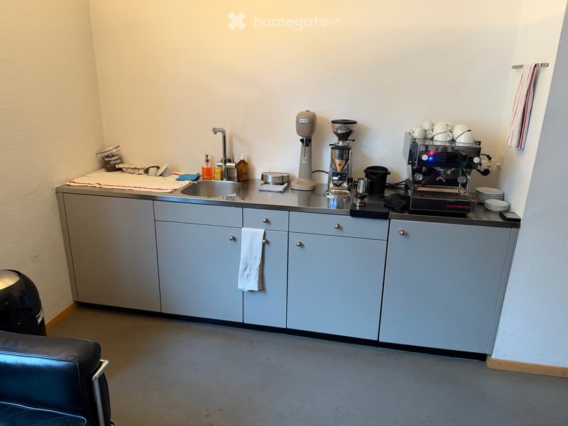 A small, modern kitchen area with a sink, coffee machine, coffee grinder, and various kitchen items on the counter. The cabinets are light blue, and there is a black leather chair in the foreground.