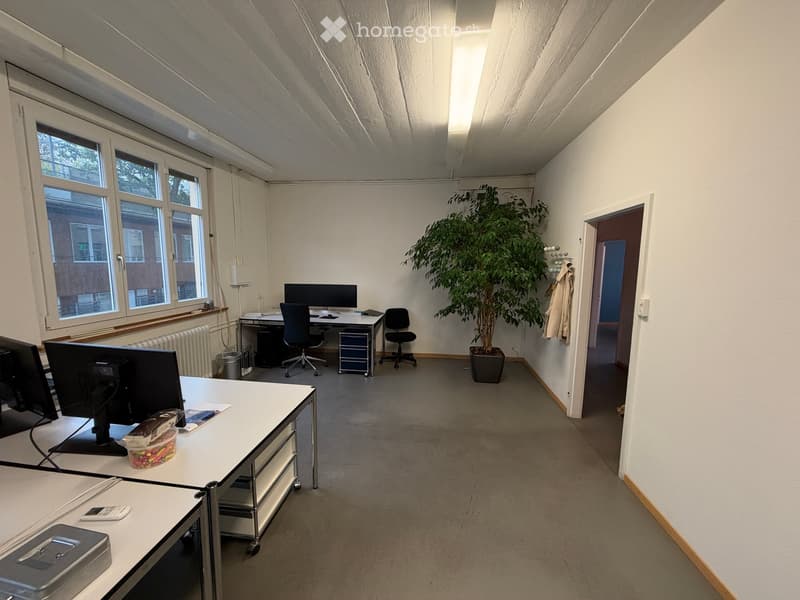 Office space with desks, chairs, computer screens, a potted plant, and a coat hanging on the wall.