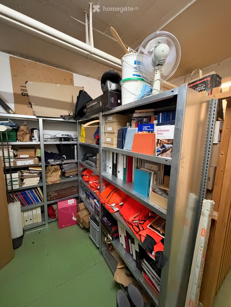 A storage room with metal shelving units filled with books, folders, boxes, and other miscellaneous items. A fan is placed on top of the shelves, and there are additional storage items scattered around the room.