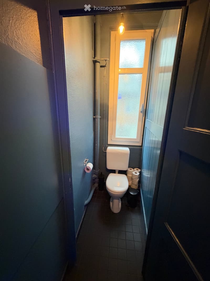 Small bathroom with a toilet, toilet paper roll on the wall, and a trash can next to the toilet. The room has dark tiled flooring and walls.