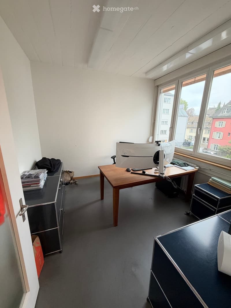 Office space with a wooden desk, computer setup, chair, and cabinet. Large windows provide natural light and a view of the city.