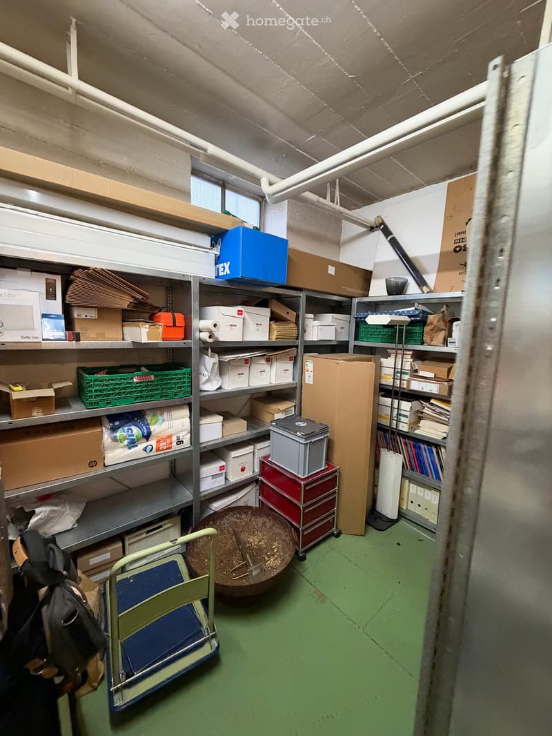 A storage room with metal shelving units filled with boxes, papers, and miscellaneous items. A green folding chair and a round wooden stool are on the floor.