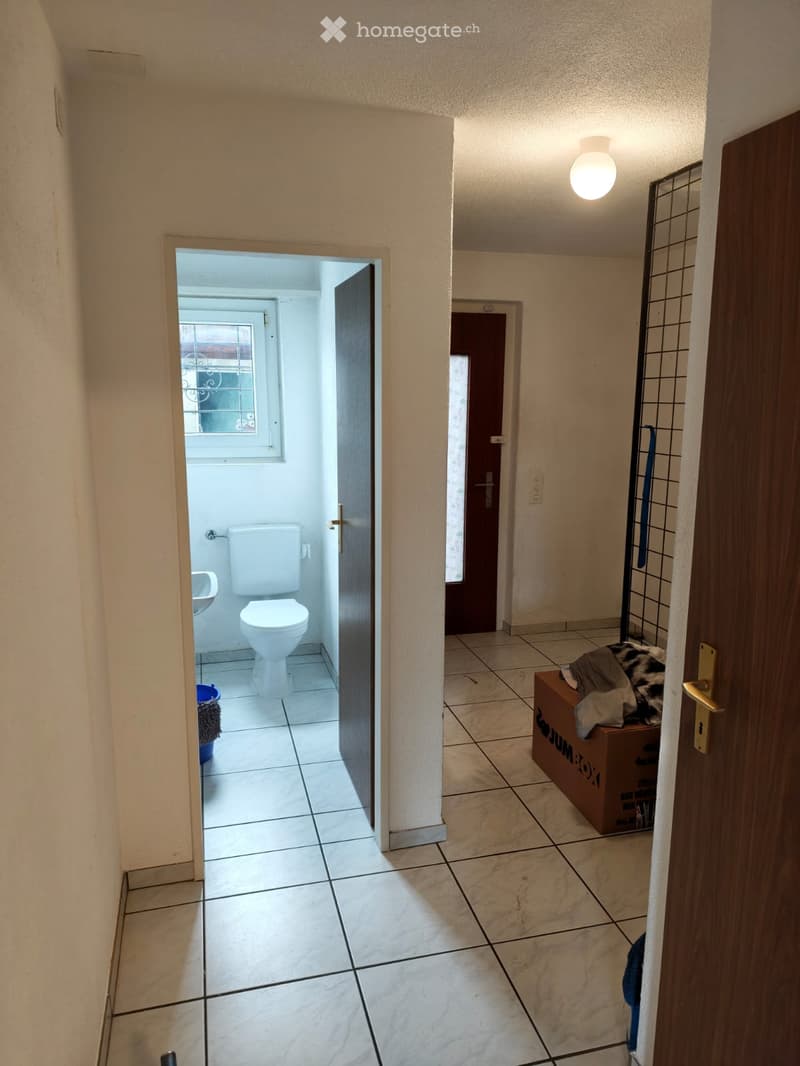 Empty hallway with tiled floor, beige walls, and a visible bathroom with a toilet and sink. A cardboard box with items is placed near the wall.