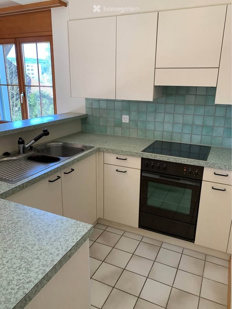 Modern kitchen with white cabinets, granite countertops, a double sink, and built-in appliances including an oven and stove. The backsplash features blue tiles, and the space is well-lit with large windows.