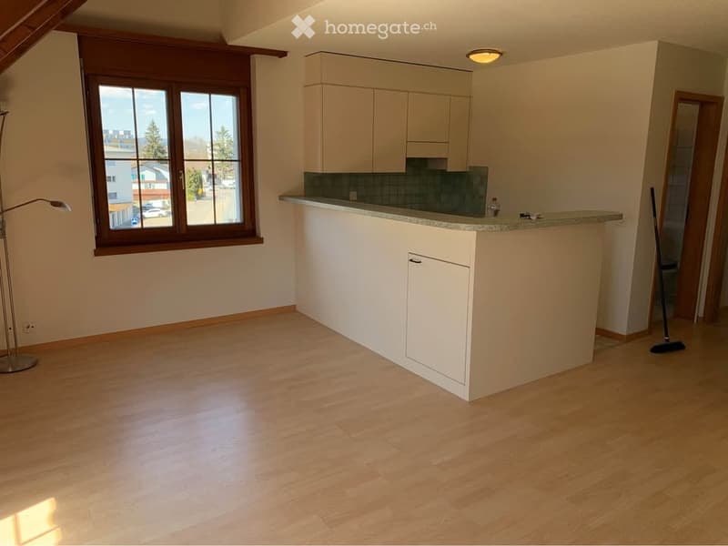 Empty kitchen with light wooden flooring, white cabinets, and a granite countertop. A large window with wooden frame provides natural light. No furniture or appliances are present.