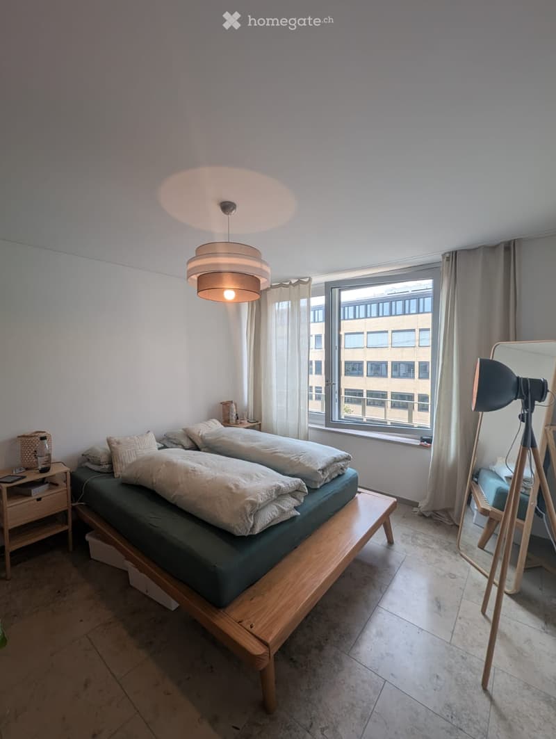 Cozy bedroom with a wooden bed, nightstand, mirror, and large window with curtains. The room features a modern pendant light and tiled flooring.