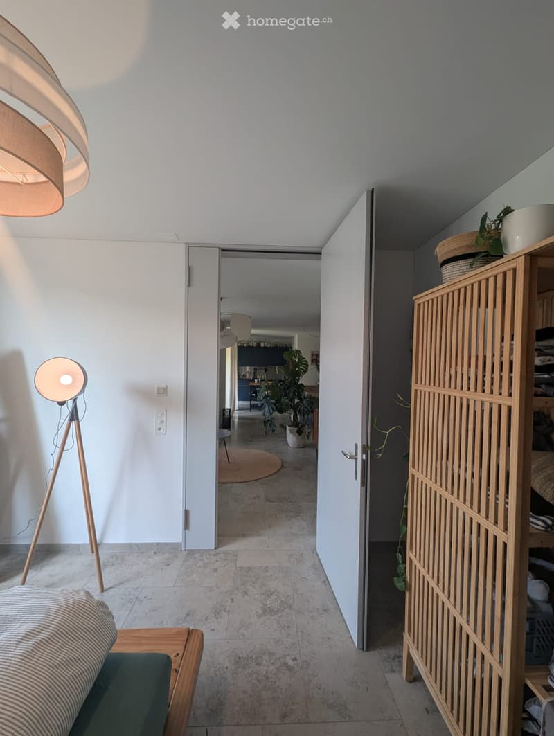 Modern hallway with a sofa, wooden shelf, and two lamps, leading to an open kitchen area with plants.
