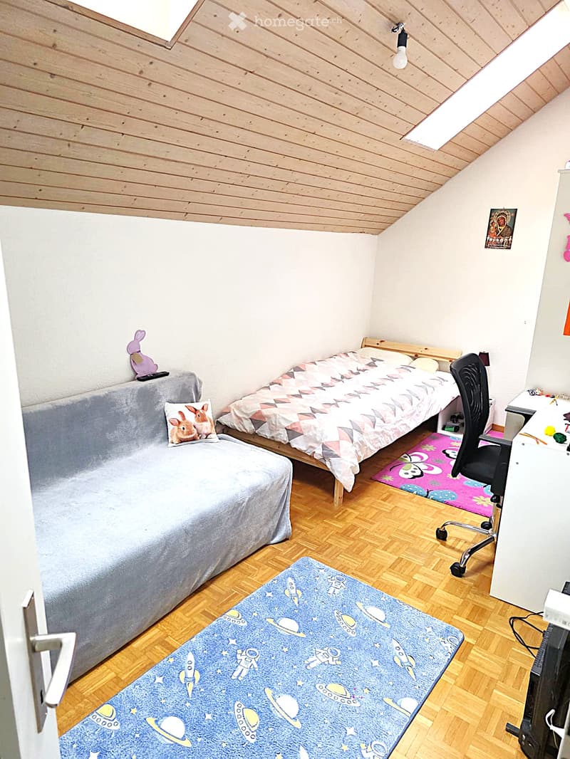 Cozy bedroom with a wooden slanted ceiling, a single bed with a colorful bedspread, a small sofa, a desk with a chair, and two rugs on the floor.