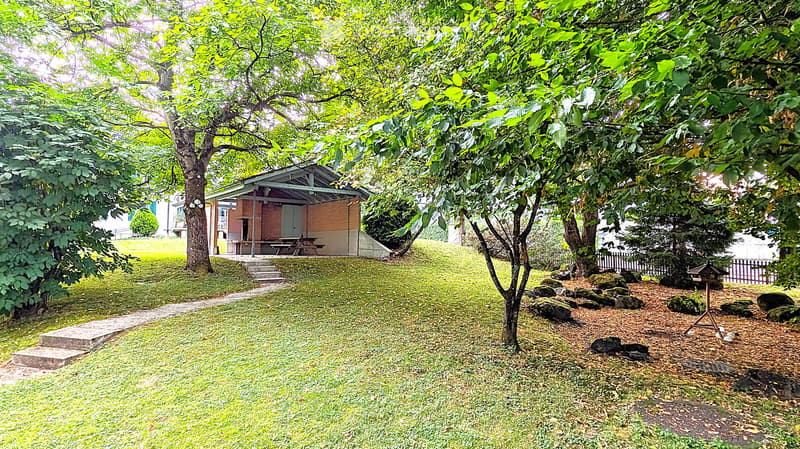 A serene garden area with a small wooden gazebo, picnic table, and lush greenery, including trees and shrubs, creating a peaceful outdoor setting.