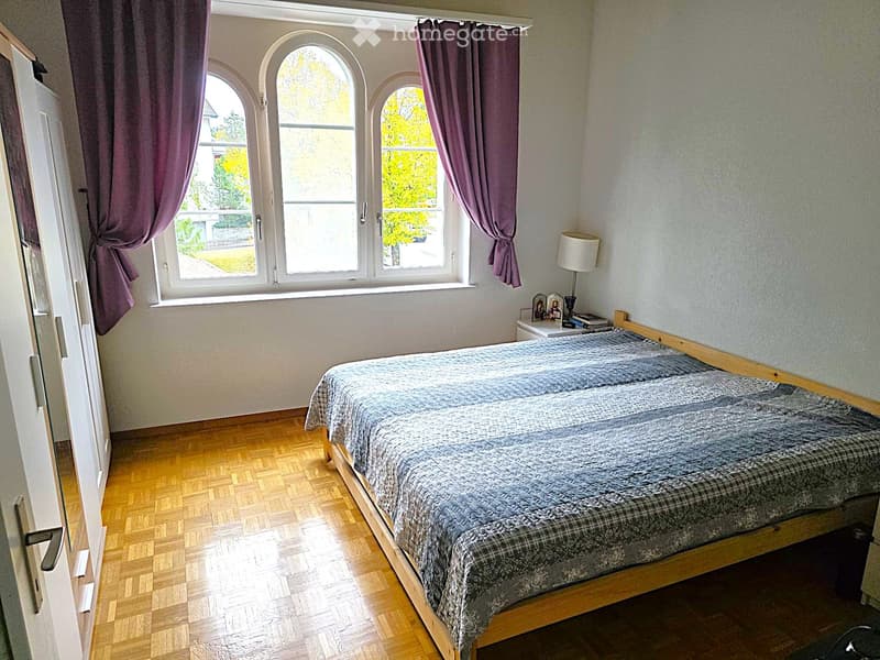 Bedroom with wooden flooring, a bed with a grey and plaid duvet, a nightstand with a lamp, and purple curtains on arched windows.