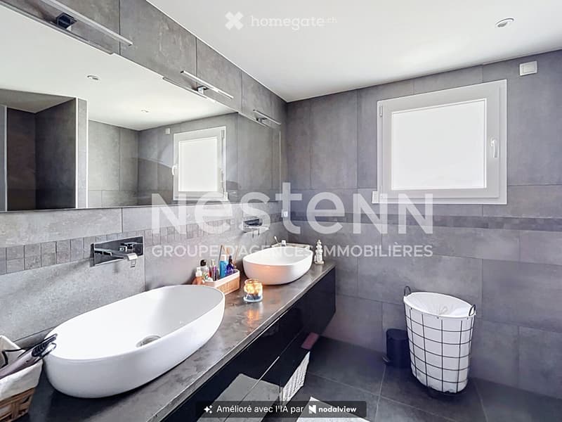 Modern bathroom with grey tiles, a large bathtub, a sink, a mirror, and a countertop with toiletries and a candle.