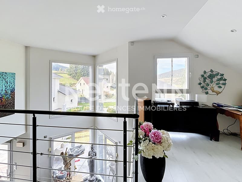 Bright, modern room with large windows offering a scenic view, featuring a table with a lamp, a vase with flowers, a decorative statue, and a mirror on the wall.