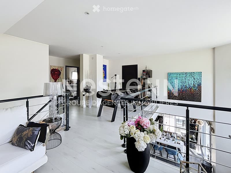 Modern, open-concept living space with white walls, light wooden flooring, and a sleek black railing. Features a white sofa, a glass coffee table, a floor lamp, a large vase with flowers, a football table, and a bookshelf with books and decorative items.