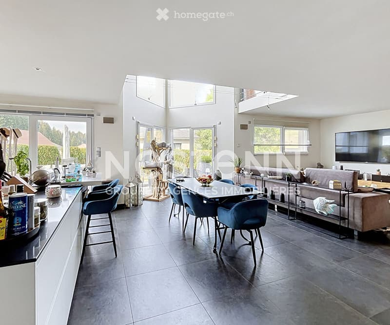 Open-plan living and dining area with modern furniture, large windows, and a kitchen counter on the left side.