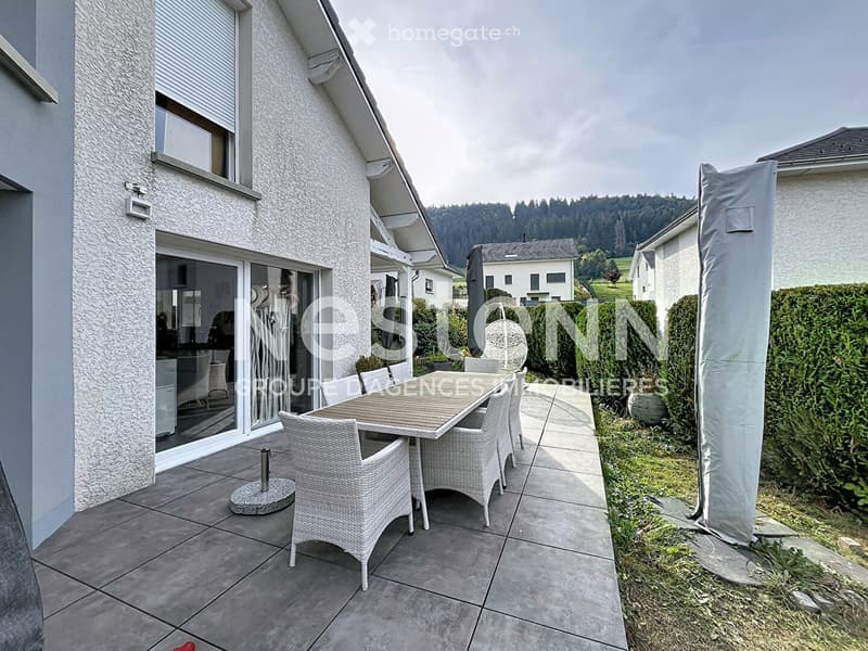 Balcony with tiled floor, outdoor dining set including a table and several wicker chairs, adjacent to a modern house with large windows and a view of the surrounding landscape.