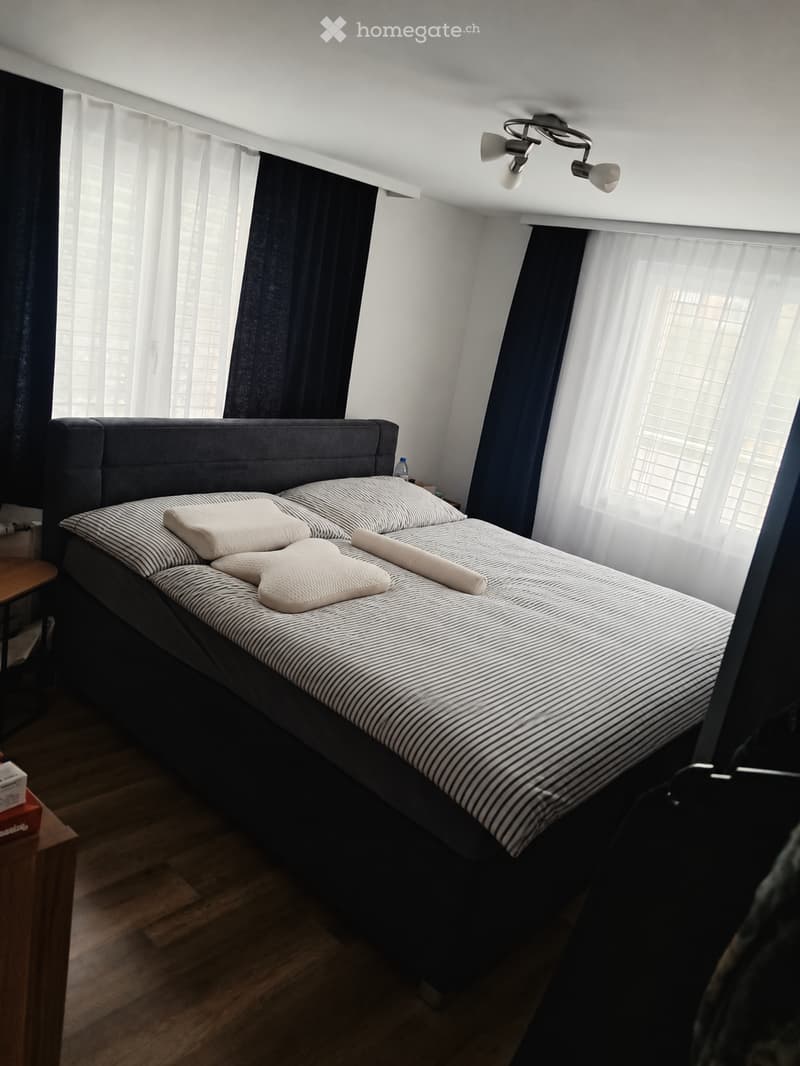 Modern bedroom with a large bed, striped bedding, and wooden flooring. Features include a nightstand, a small table, and large windows with dark curtains.