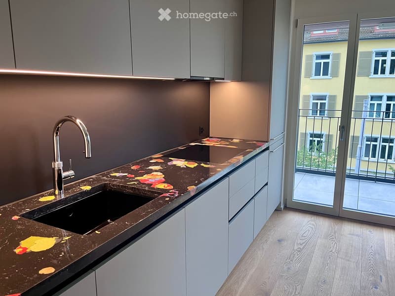 Modern kitchen with a dark countertop featuring a leaf design, a sleek faucet, and white cabinets. A sliding glass door opens to a balcony with a view of a neighboring building.