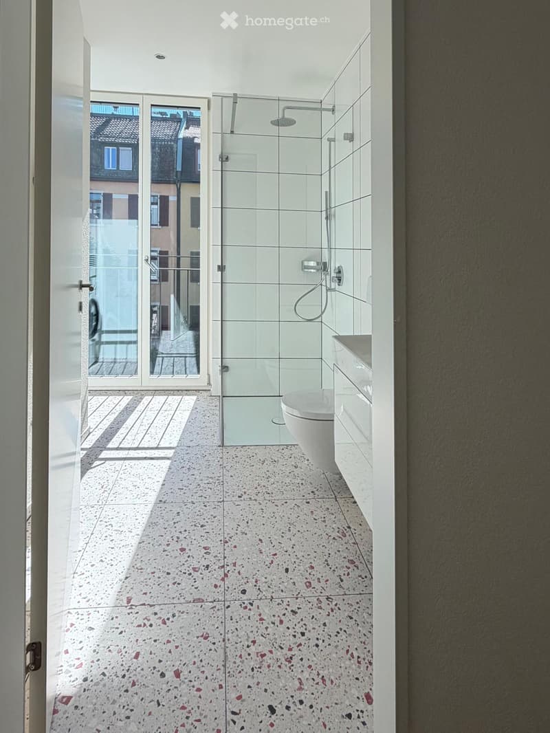 Modern bathroom with white tiles, a glass-enclosed shower, a toilet, and a sink. The floor features a speckled tile design.