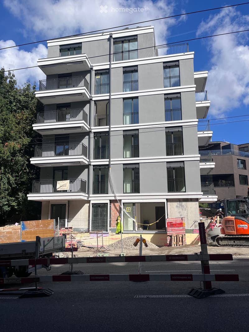 Modern multi-story residential building with large windows and balconies, under construction with visible barriers and construction equipment.