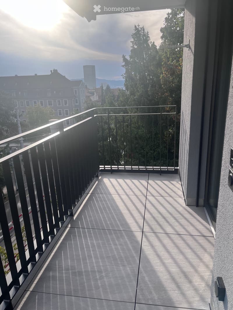 Balcony with modern black metal railing, tiled floor, and cityscape view including trees and buildings.