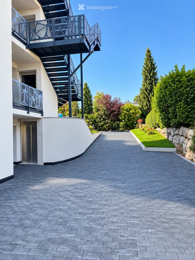 Brick-paved pathway leading to a modern building with balconies and decorative railings, surrounded by well-maintained greenery and trees.