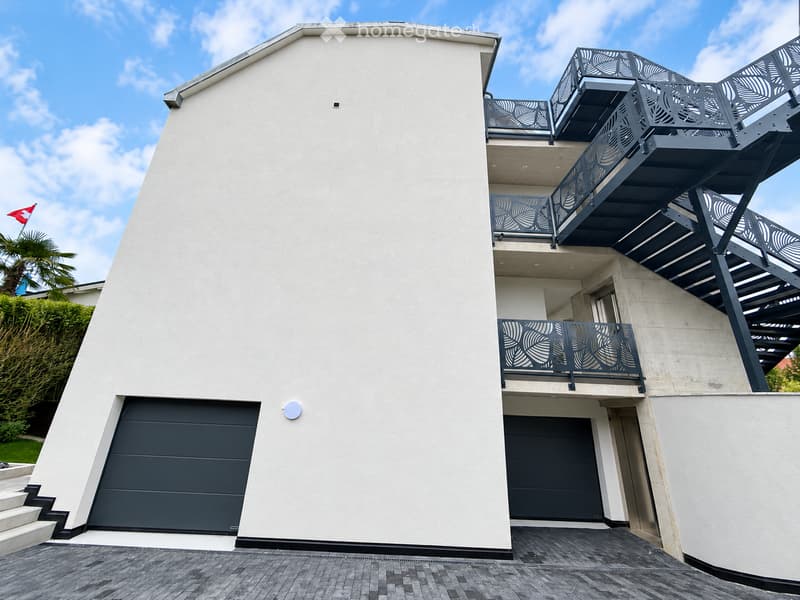 Modern two-story house with white exterior, dark grey garage doors, and multiple balconies with decorative railings.