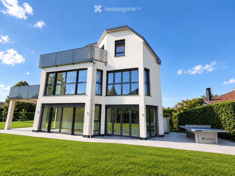 Modern two-story house with white exterior, large windows, and a glass balcony. The house features a well-maintained lawn and a ping pong table in the garden area.