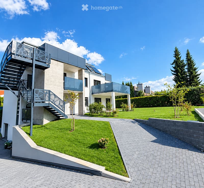 Modern two-story house with a raised ground floor, featuring a balcony, staircase, and well-maintained garden with a lawn and pathway.