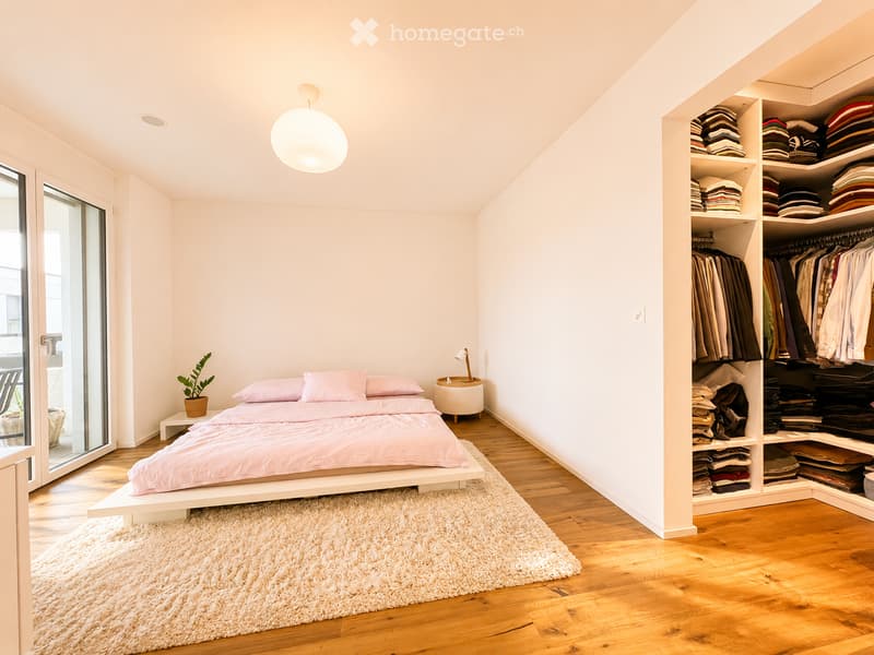 Bright bedroom with a white bed, pink bedding, a built-in wardrobe, a small nightstand with a lamp, and a potted plant near the window.