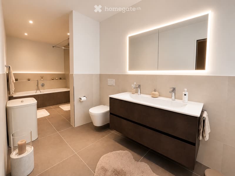 Modern bathroom with a large illuminated mirror, double sink, toilet, bathtub, and decorative candles.